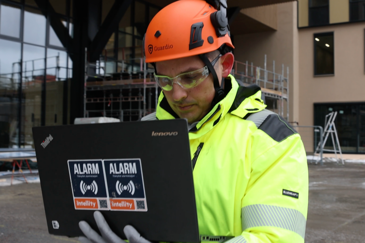 An engineer wearing a yellow high-visibility work uniform and a safety helmet, standing on a construction site while holding a laptop. The engineer is focused on the screen, likely analyzing data or managing project tasks in a rugged outdoor environment.